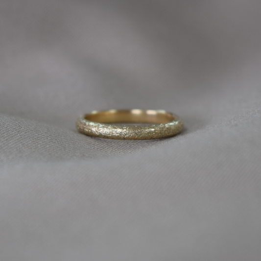 A thin textured gold band ring resting on a soft, draped gray fabric background.