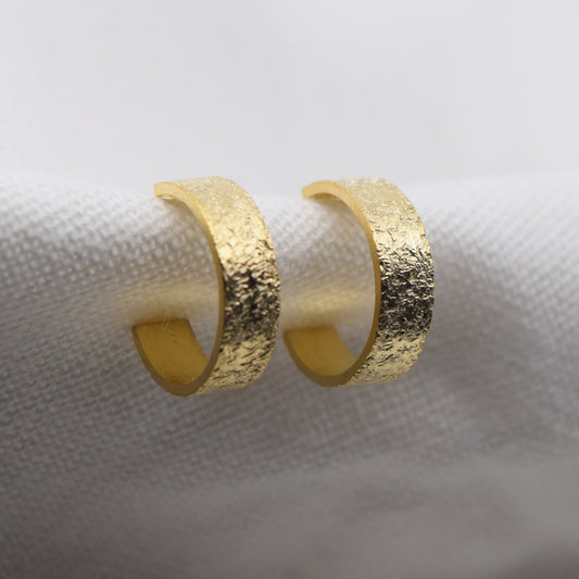 A pair of gold-colored mini textured hoop earrings placed on a white fabric surface.