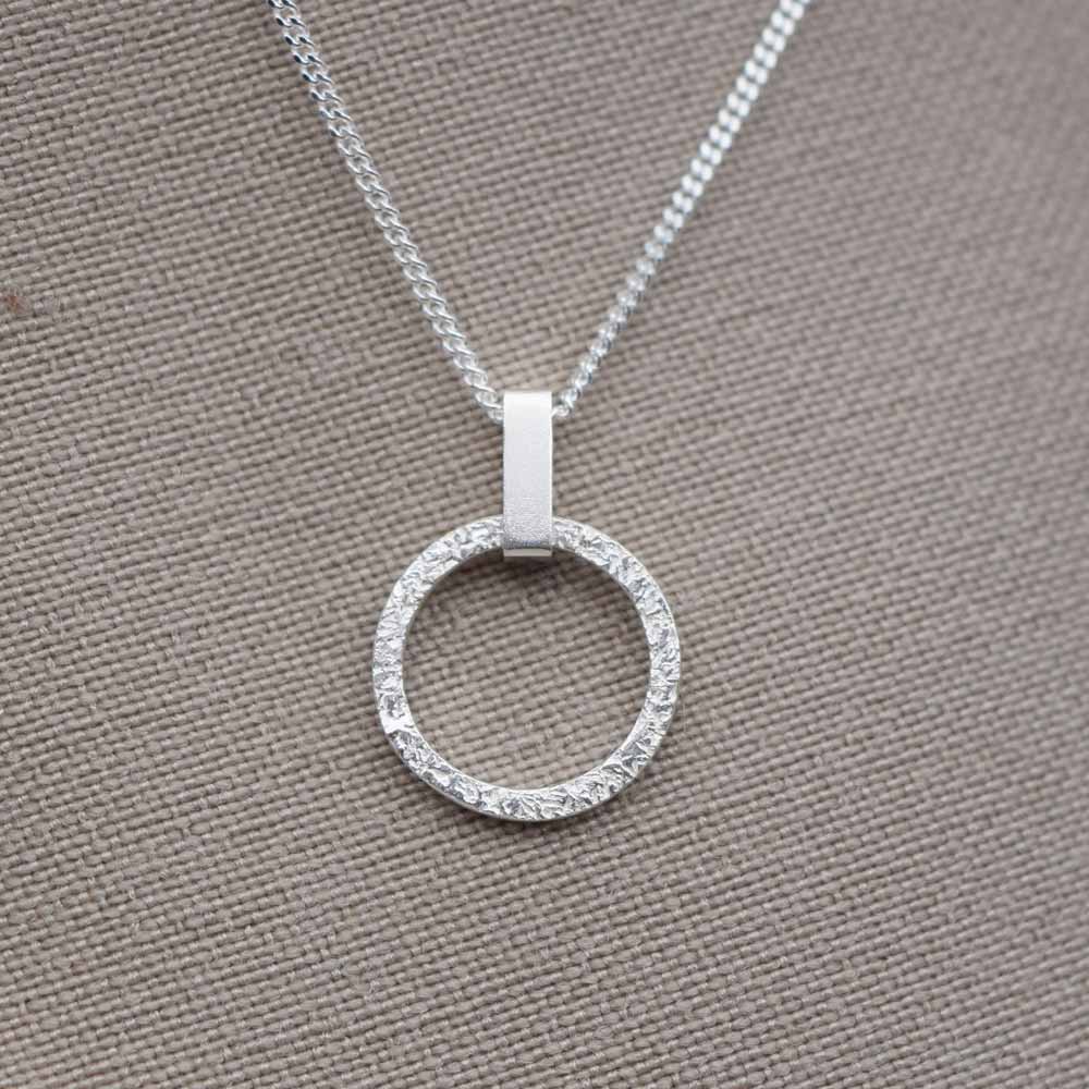 A small, round, textured silver pendant with a mini rectangle link on a chain, displayed against a fabric background.