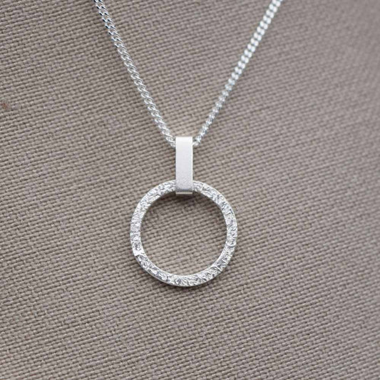 A small, round, textured silver pendant with a mini rectangle link on a chain, displayed against a fabric background.