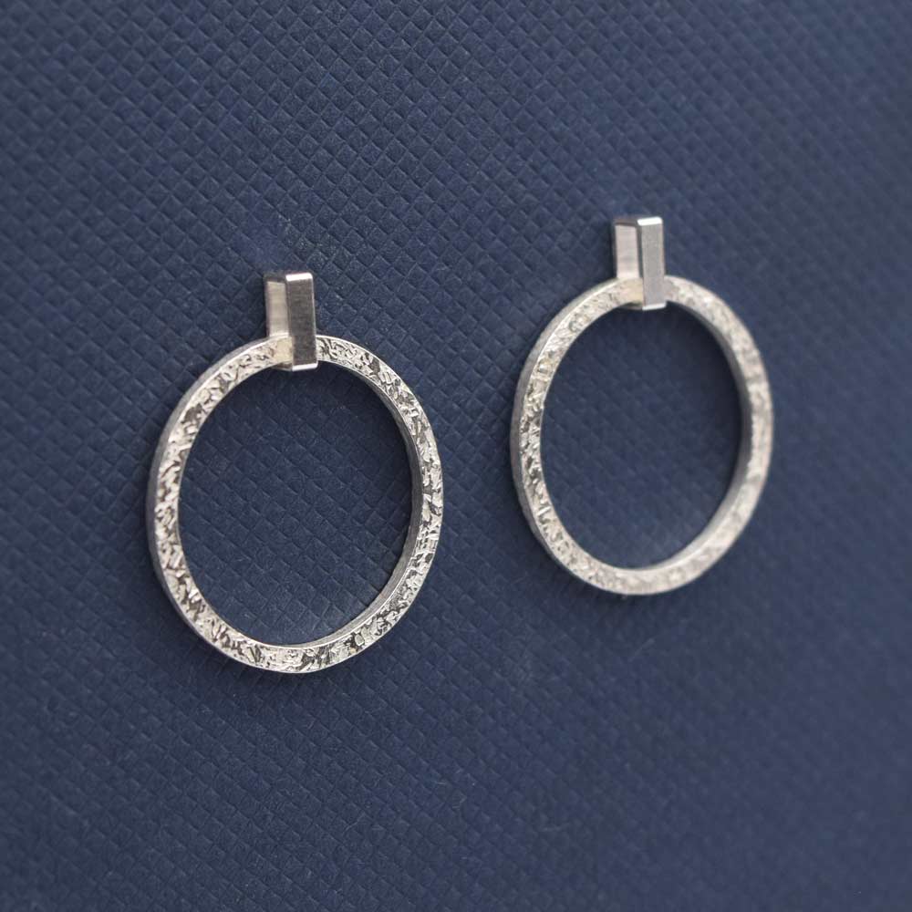 A pair of silver textured drop earrings with a movable loop, displayed against a blue background.