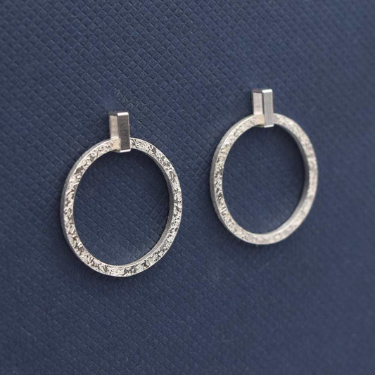 A pair of silver textured drop earrings with a movable loop, displayed against a blue background.